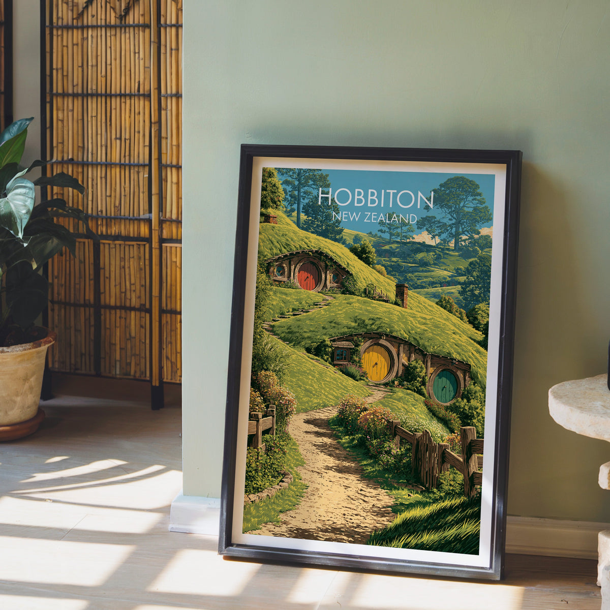 Waikato farmland artwork in a black frame on a pale wood floor by a sage-green wall and bamboo screen
