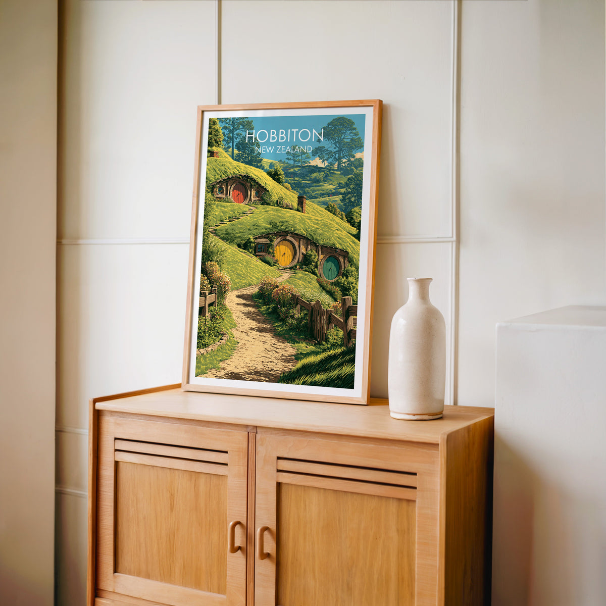 Green pasture and yellow door print in an oak frame on a light oak cabinet by a cream panelled wall