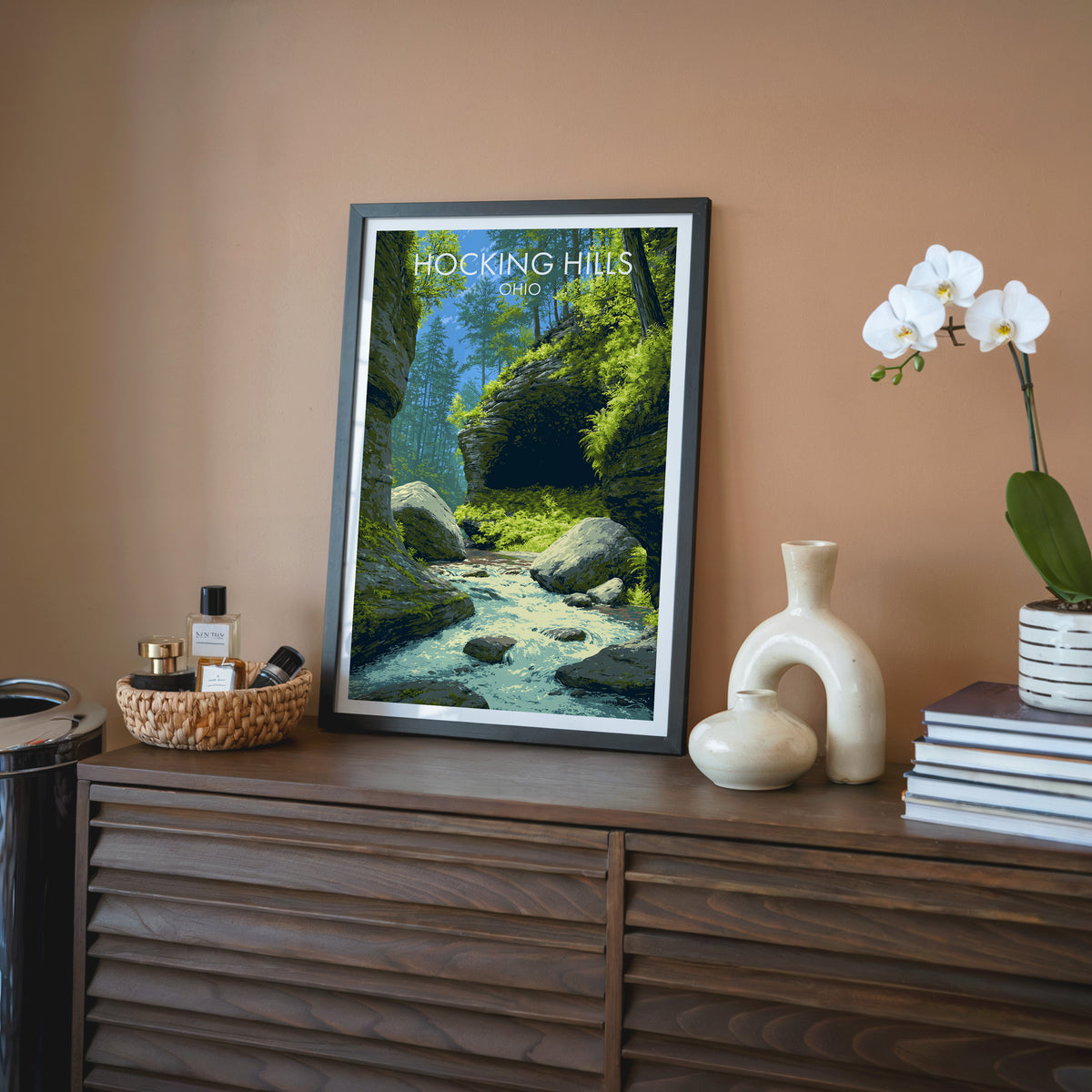 Forest gorge wall art in a black frame on a walnut sideboard with terracotta wall, orchid and cream vase