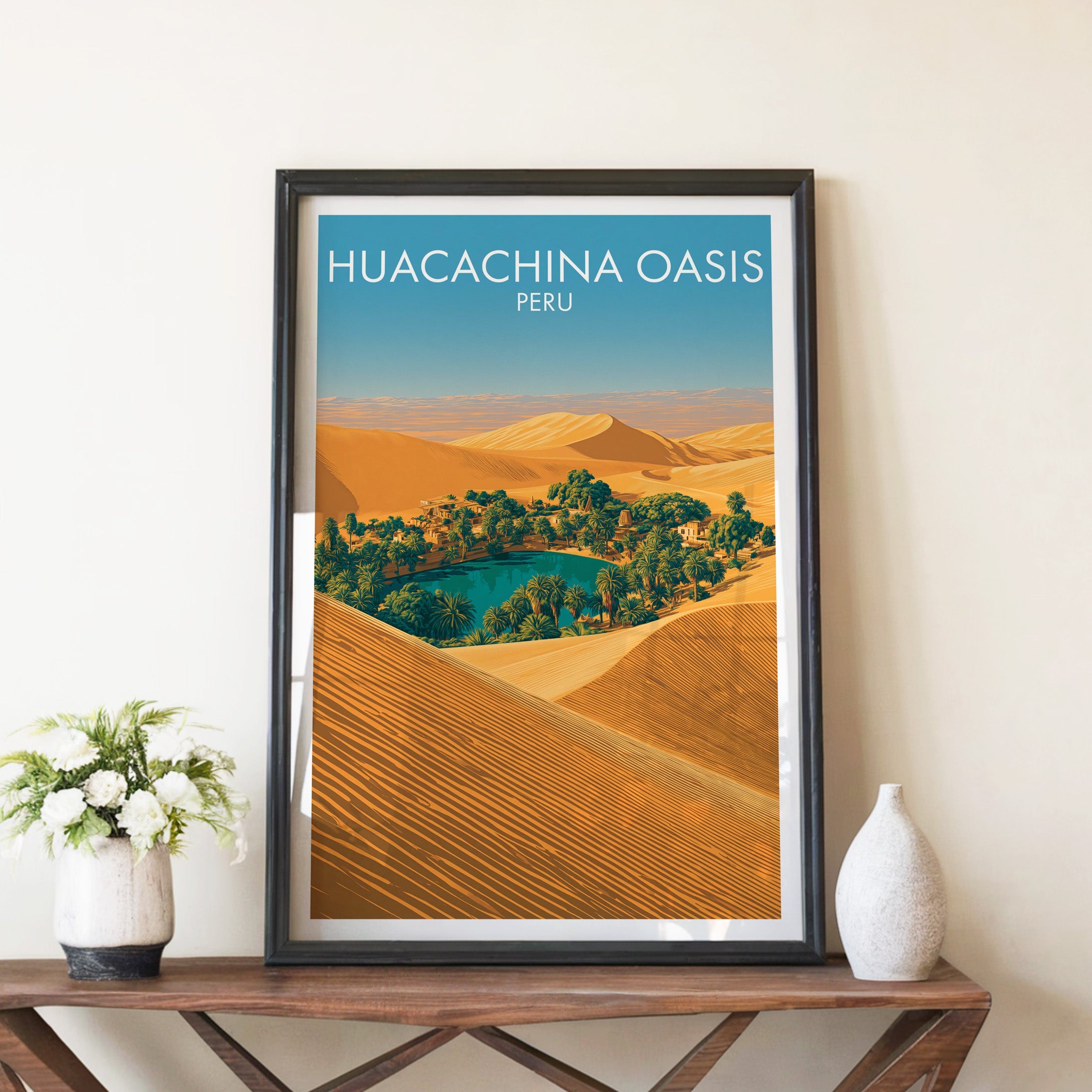 Desert oasis print in a black frame on a brown console table with white flowers in a ceramic vase
