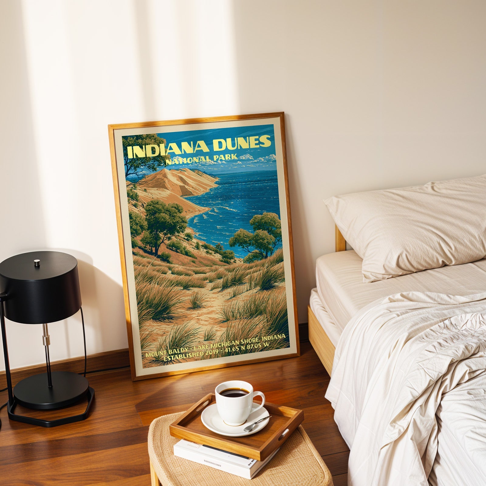 Mount Baldy dune poster in a gold frame on the bedroom floor beside a made bed and a black table lamp