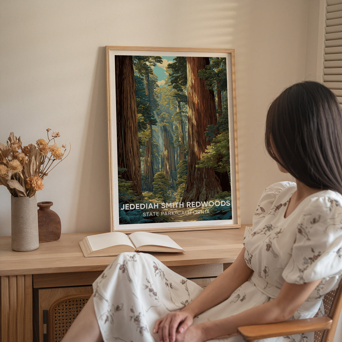 A woman in a white dress sits beside the oak-framed Jedediah Smith Redwoods print on a sideboard with dried flowers.