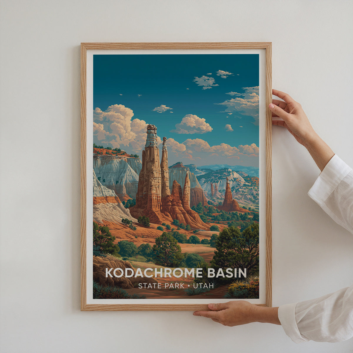 Two hands holding the oak-framed Kodachrome Basin print flat against a plain white wall.