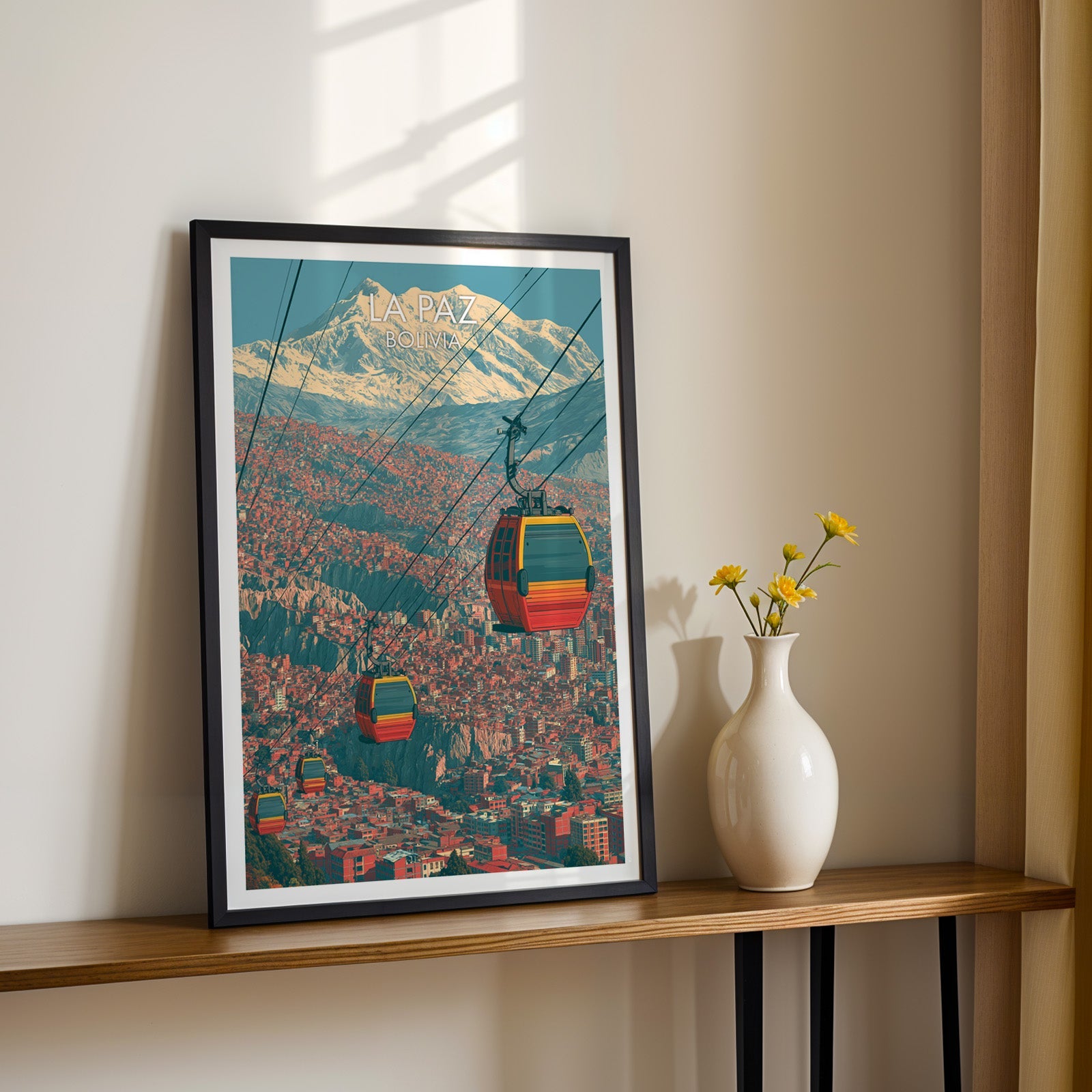 Bolivian cable car print in a black frame on a slim wood shelf with yellow daisies in a white vase, sunlit cream wall