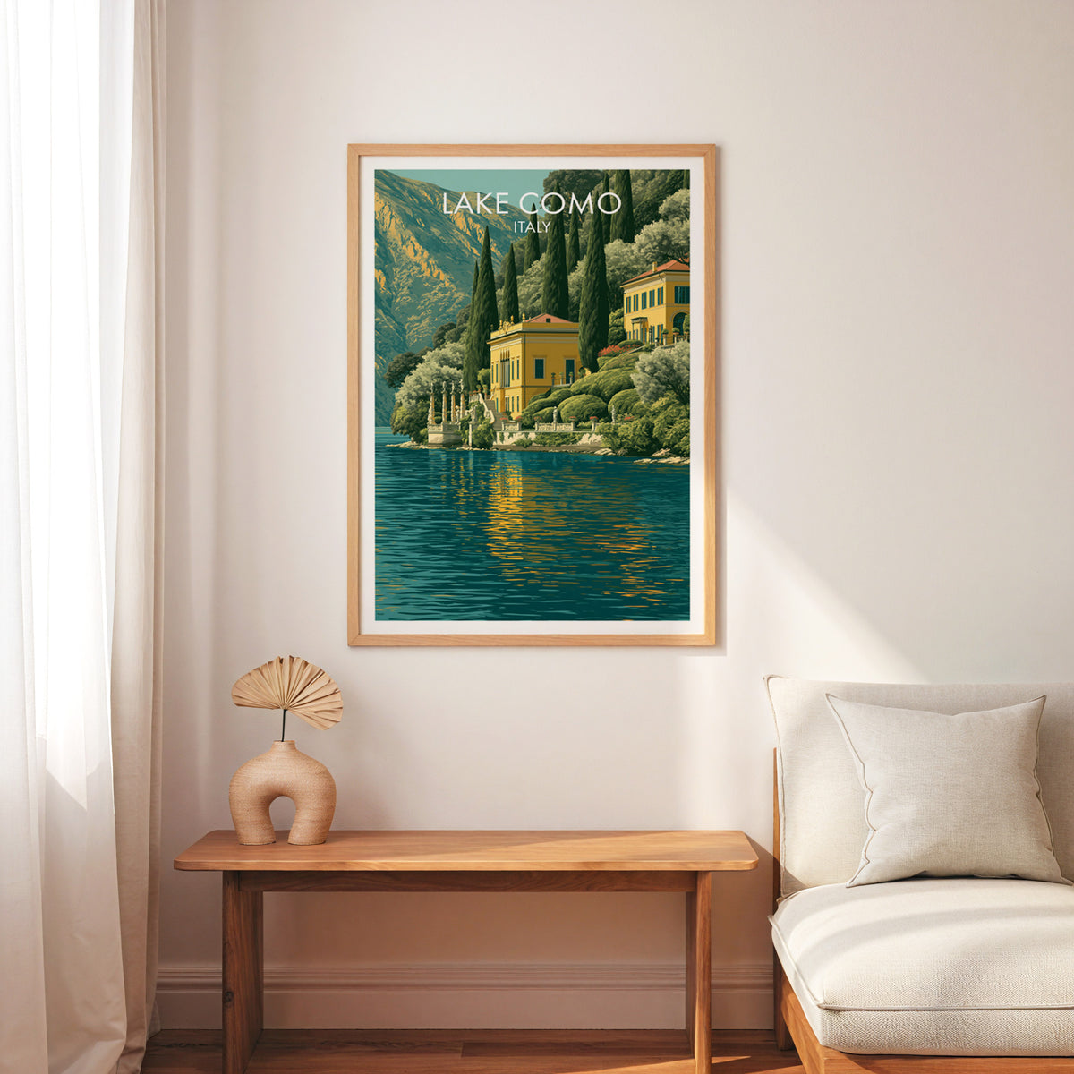 Lake Como travel wall art in an oak frame above a slim wooden bench, cream armchair and a palm leaf fan