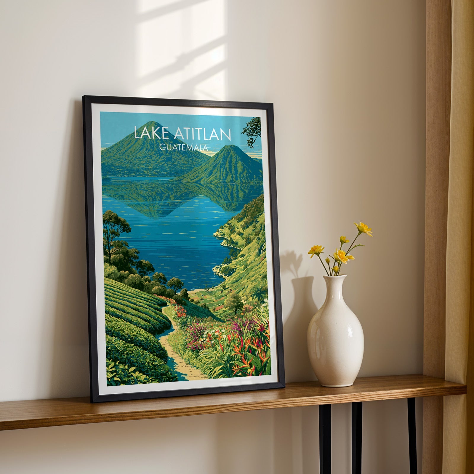Guatemala highland landscape in a black frame on a narrow wood shelf, yellow daisies beside it on a sunlit cream wall