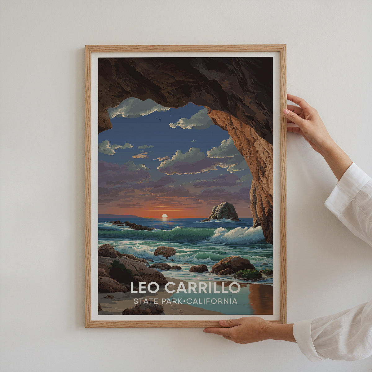 Two hands hold the oak-framed Leo Carrillo travel poster flat against a plain white wall.
