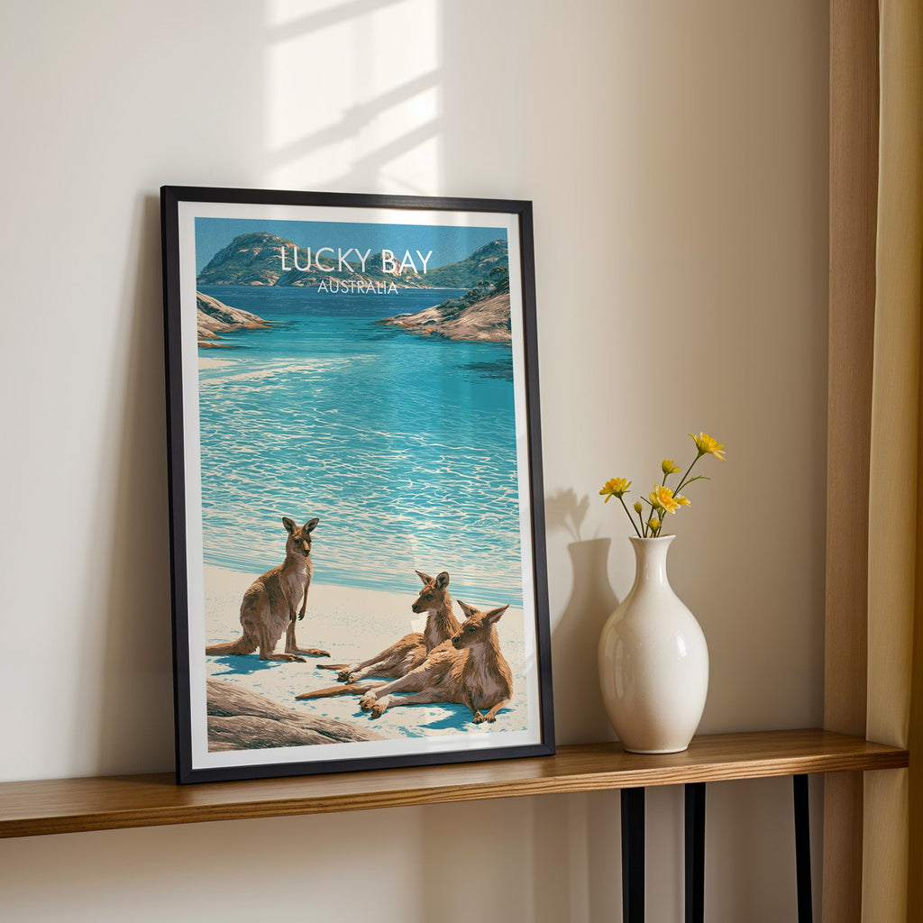 Turquoise Lucky Bay artwork in a black frame on a wood shelf with yellow daisies on a sunlit cream wall