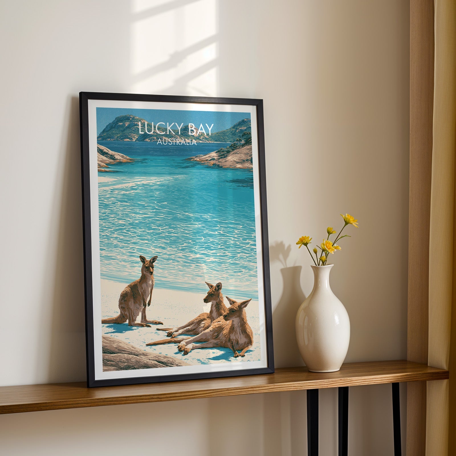 Turquoise Lucky Bay artwork in a black frame on a wood shelf with yellow daisies on a sunlit cream wall