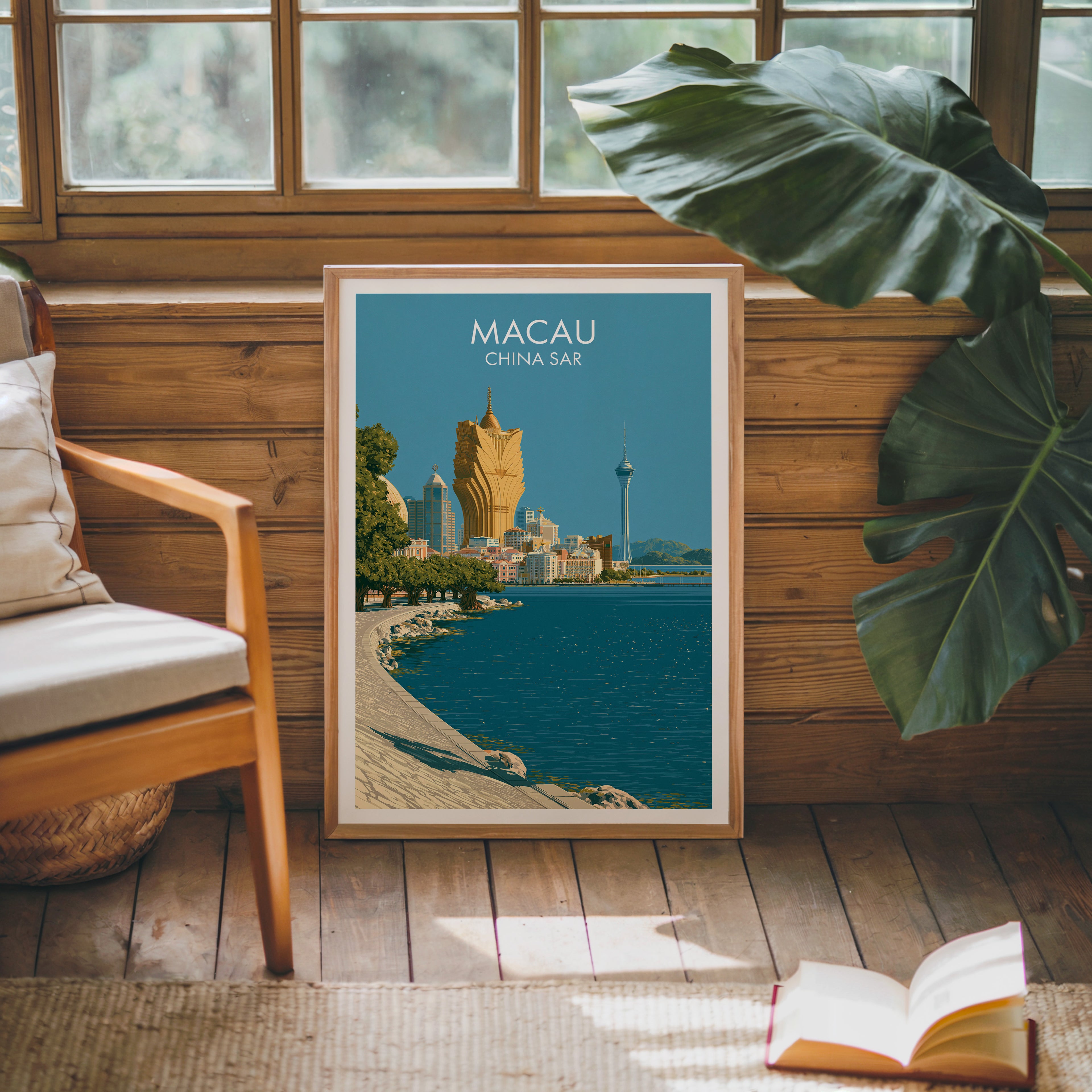 Framed Macau China poster leaning against a timber cabin wall with monstera leaves and an open book