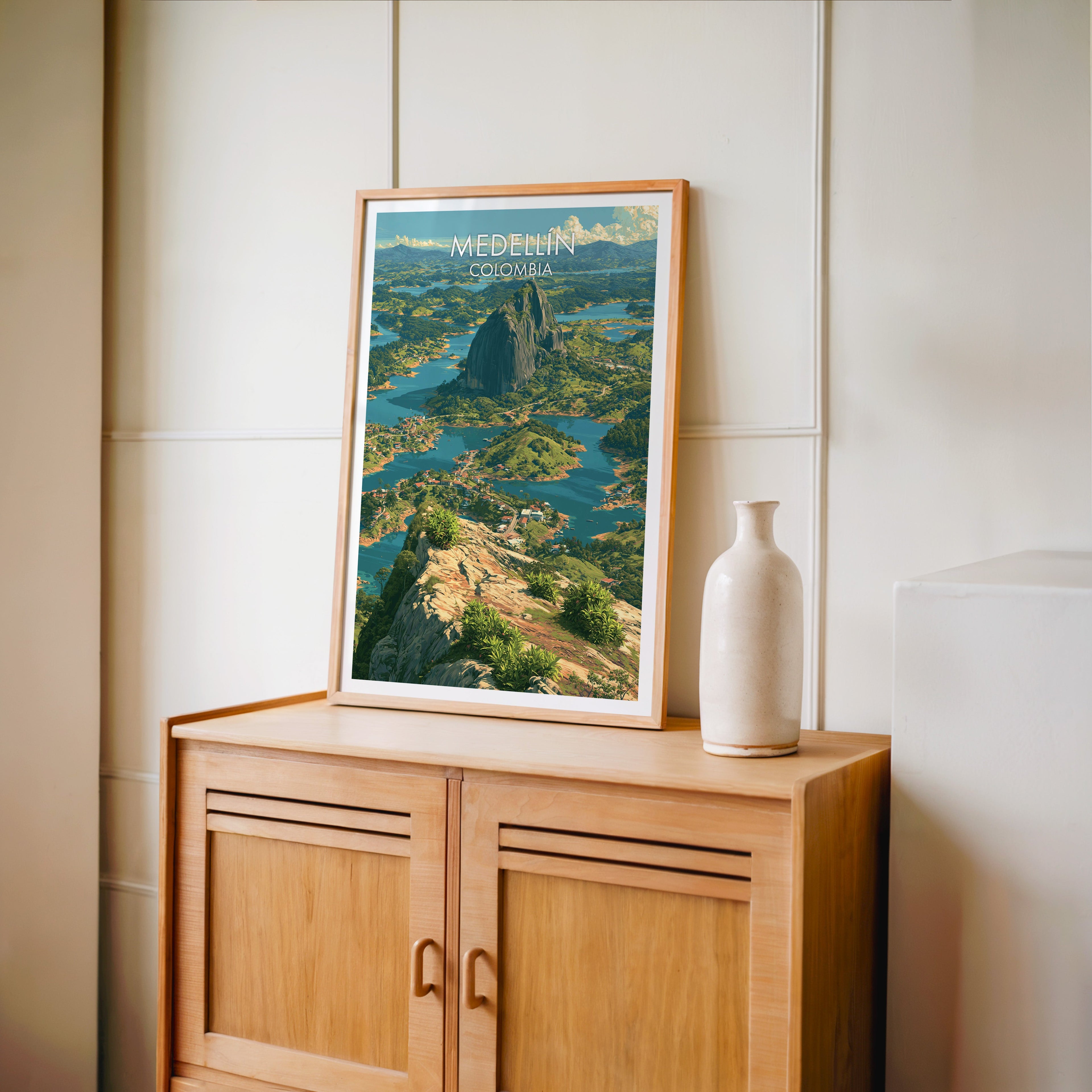 Medellin travel poster in an oak frame standing on a light oak cabinet against a cream panelled wall with a tall vase