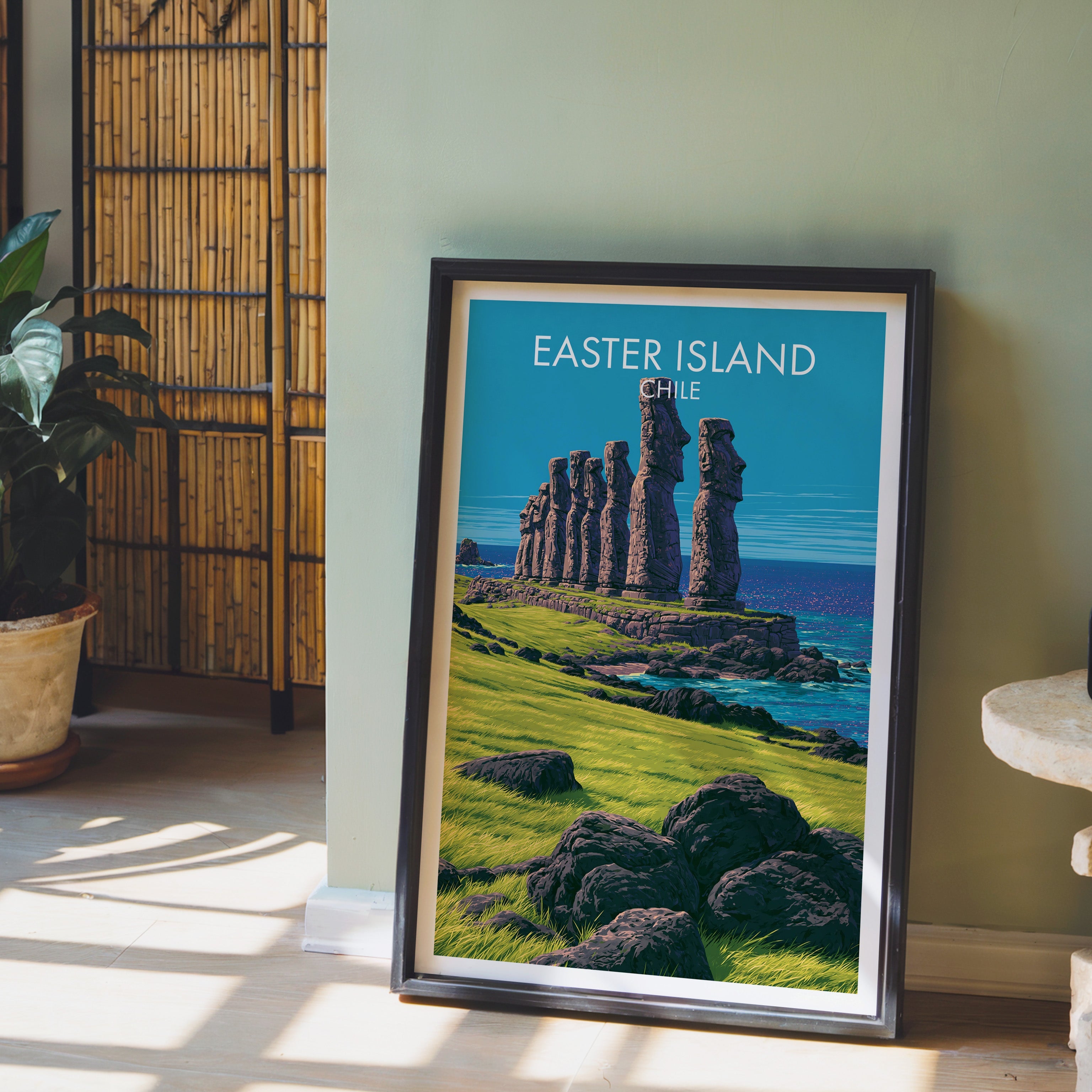 Easter Island coast print in a dark brown frame on a light wood floor by a sage wall, bamboo screen and potted plant