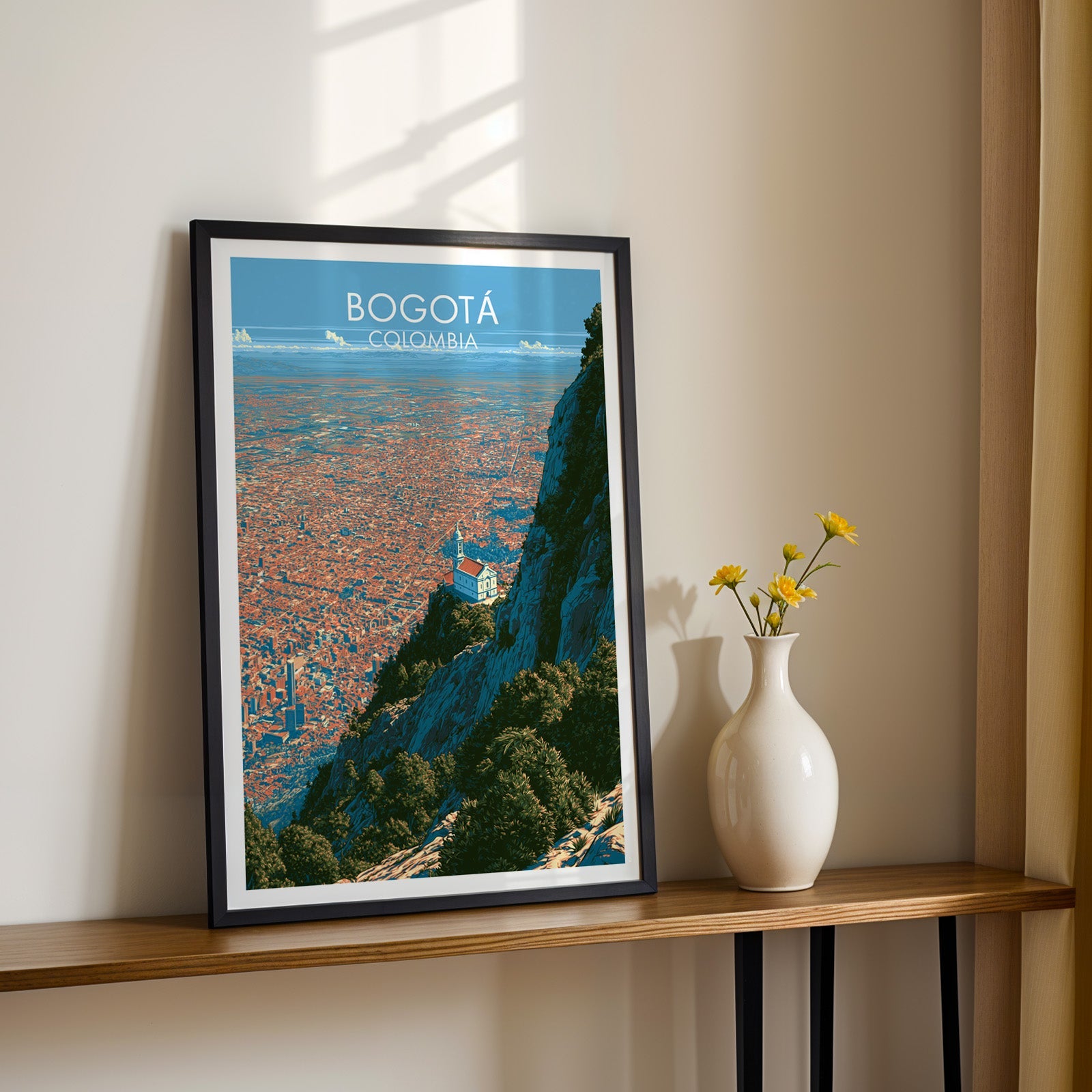 Monserrate sanctuary print in a black frame on a slim wood shelf with yellow daisies in a white vase, cream wall