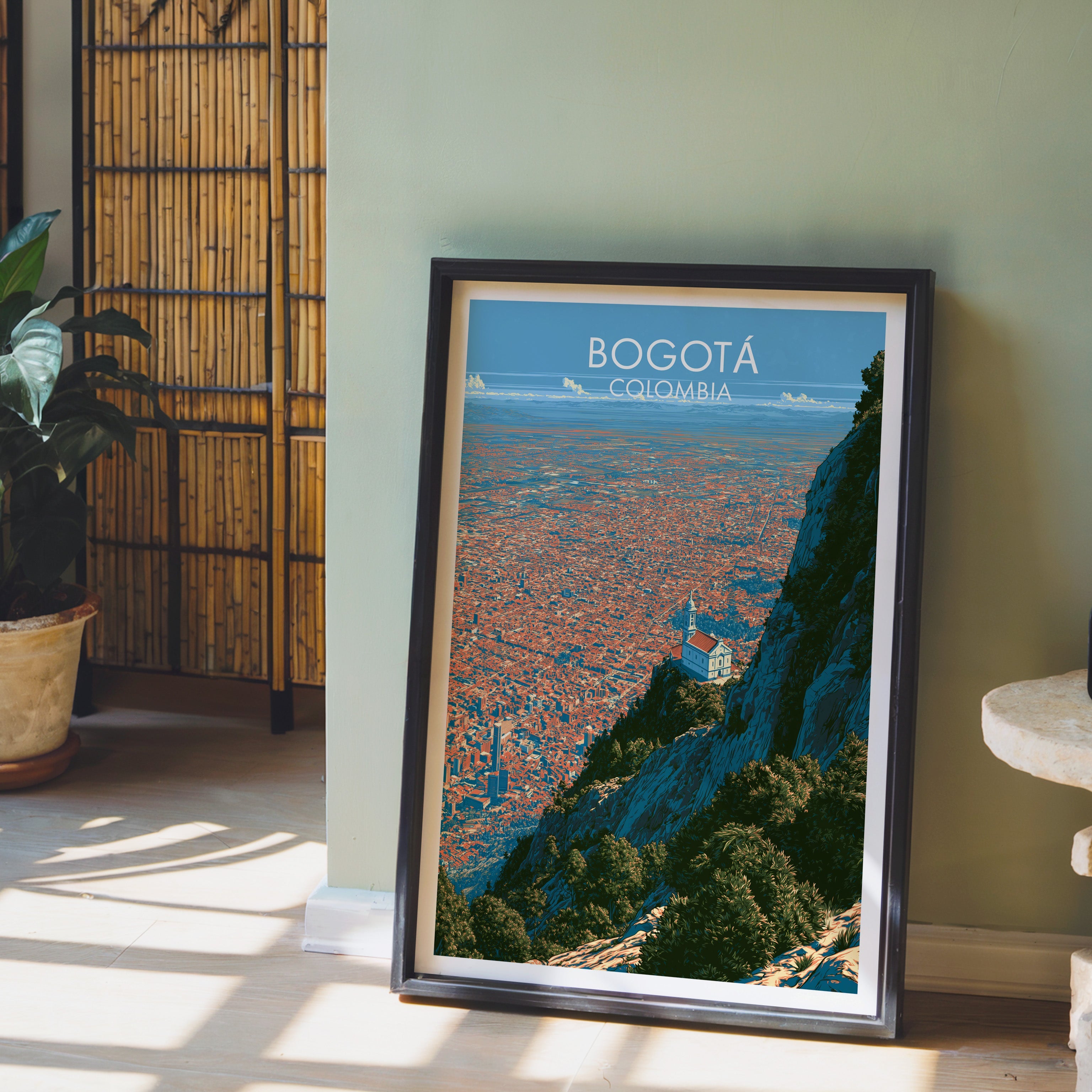 Bogota cityscape art in a dark brown frame on pale wood flooring by a sage green wall, bamboo screen and clay pot