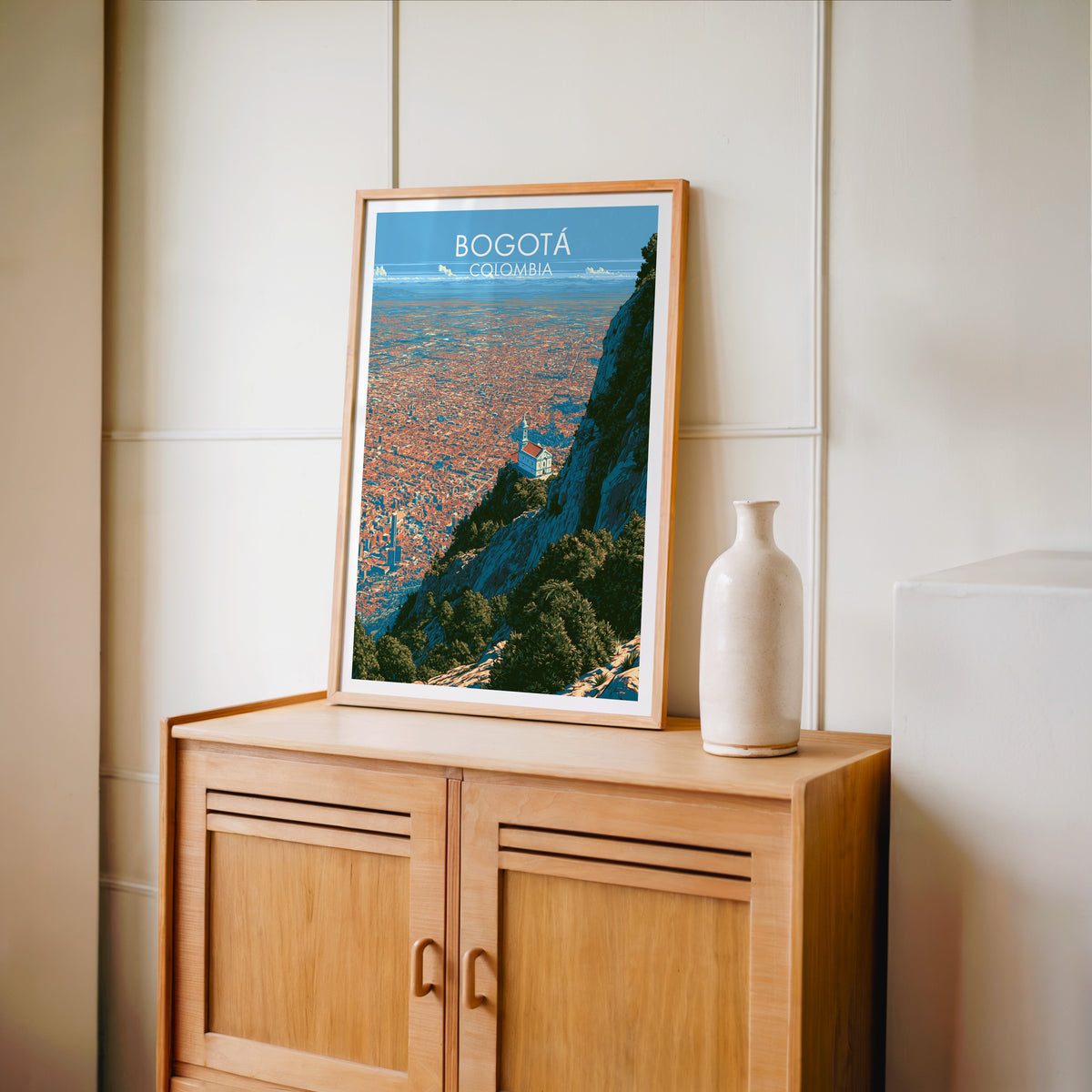 Bogota travel poster in an oak frame standing on a light oak cabinet against a cream panelled wall with a tall vase