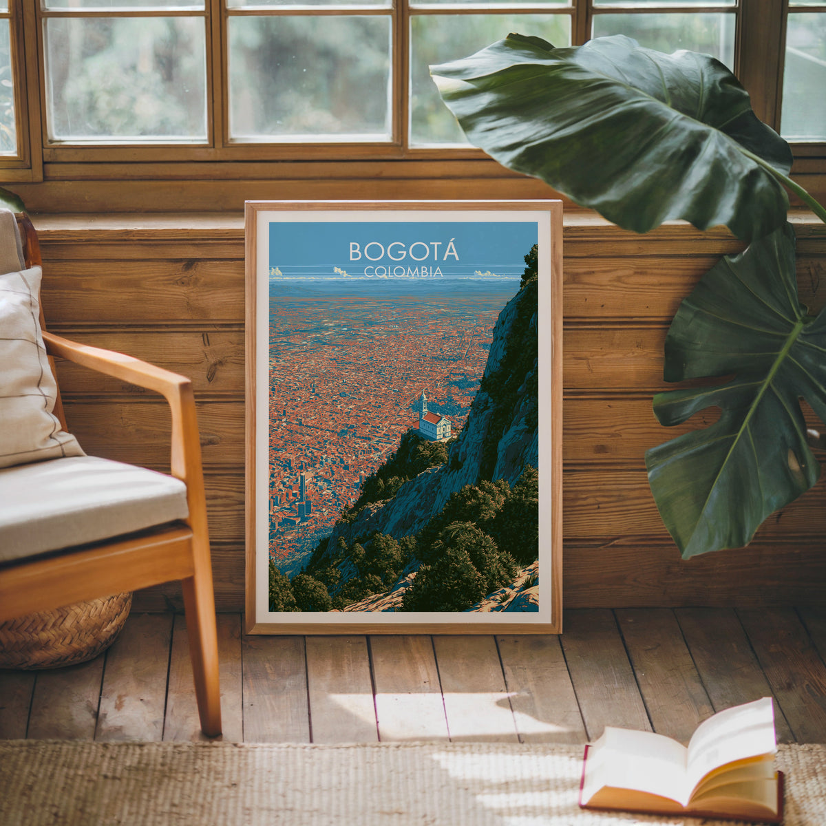 Hilltop church print in an oak frame on a pine cabin wall beside a cream cushioned chair, monstera leaf and open book