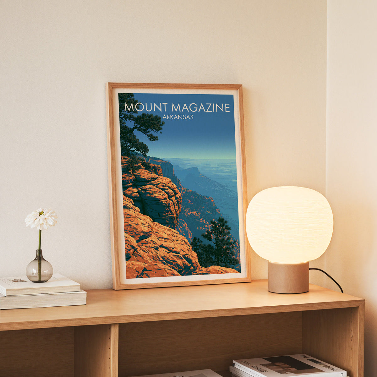 Ozark escarpment print in a slim oak frame standing on an oak console beside white daisies in a glass jar