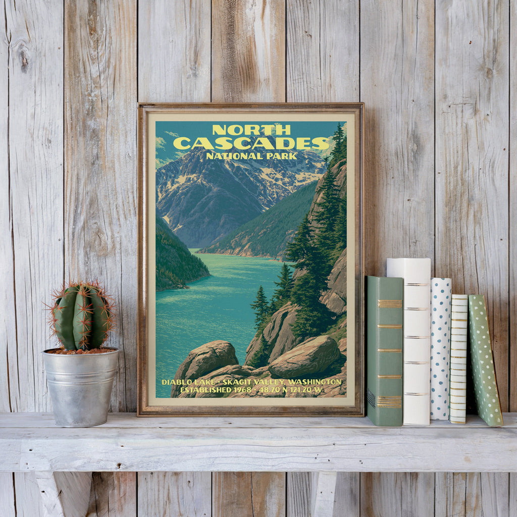 North Cascades vintage poster in a slim gold frame on a whitewashed plank wall beside a cactus and pastel books