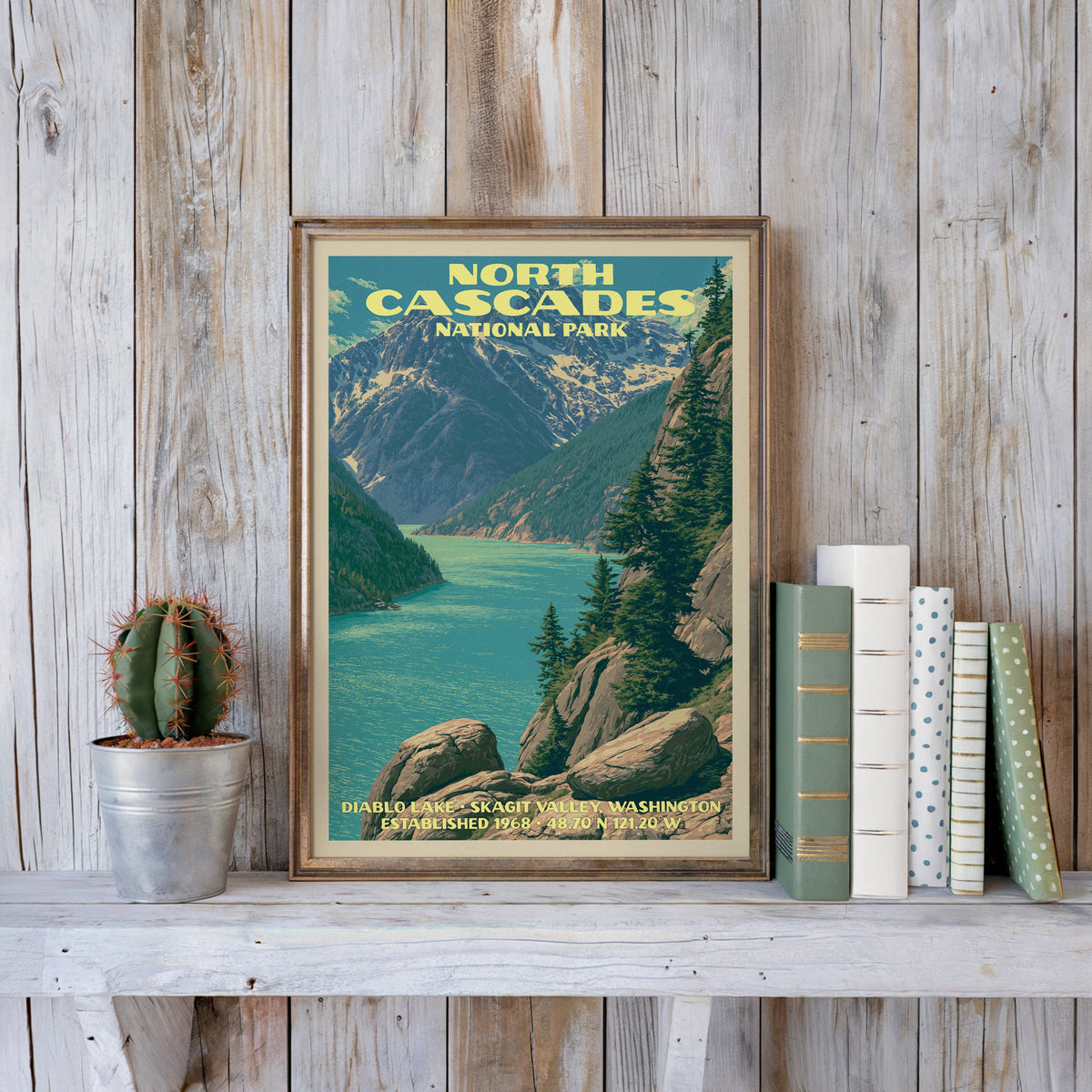 North Cascades vintage poster in a slim gold frame on a whitewashed plank wall beside a cactus and pastel books