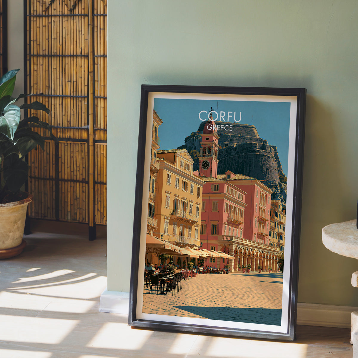 Corfu town poster, dark frame leaning on a sage-green wall beside a bamboo screen and a peace lily