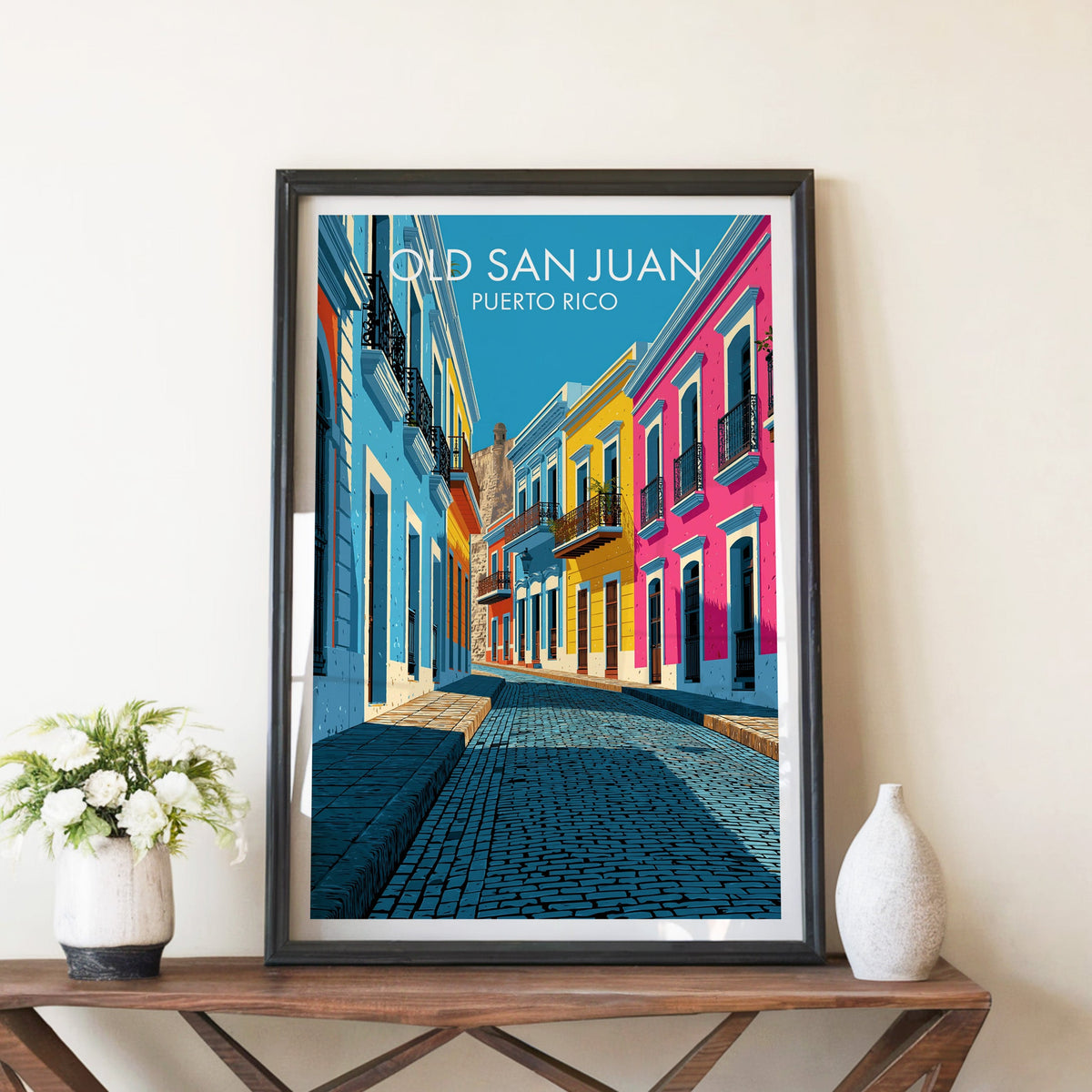 Pink and blue facades of Old San Juan in a black frame on a walnut console with white peonies and a white vase