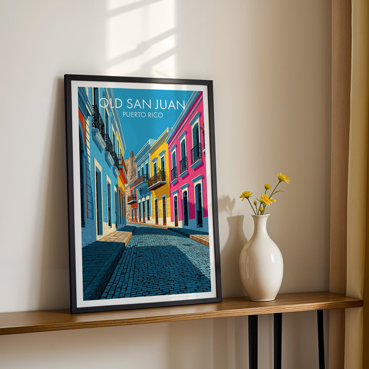 Cobbled street in Puerto Rico in a black frame on a narrow wood shelf with yellow daisies and a sunlit cream wall