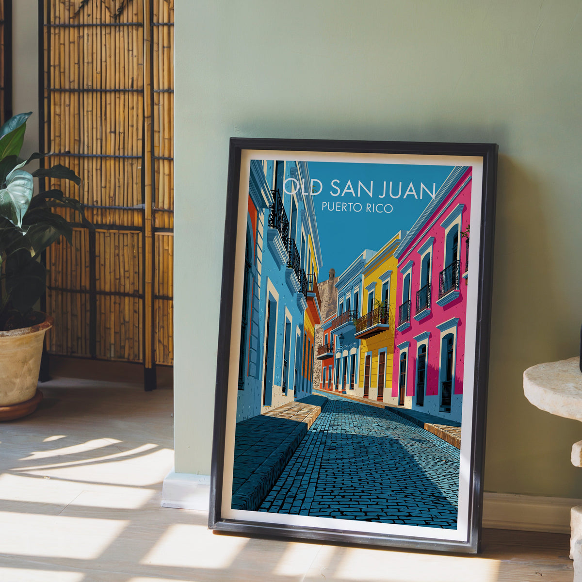 Iron balcony detail print in a black frame leaning on pale wood floor by a sage-green wall with a bamboo screen
