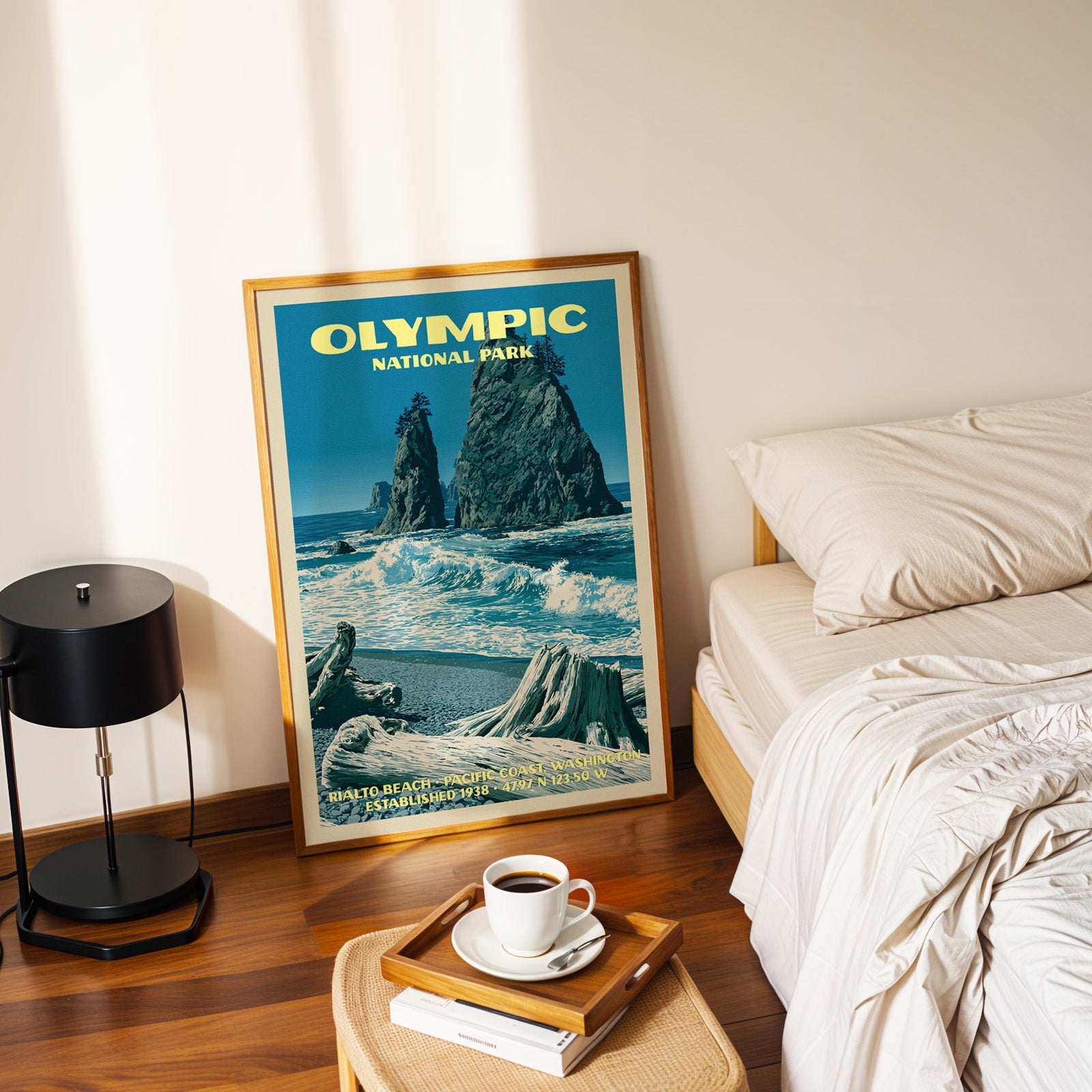 Rialto Beach Olympic poster in a gold frame on the floor beside a made bed and a black table lamp