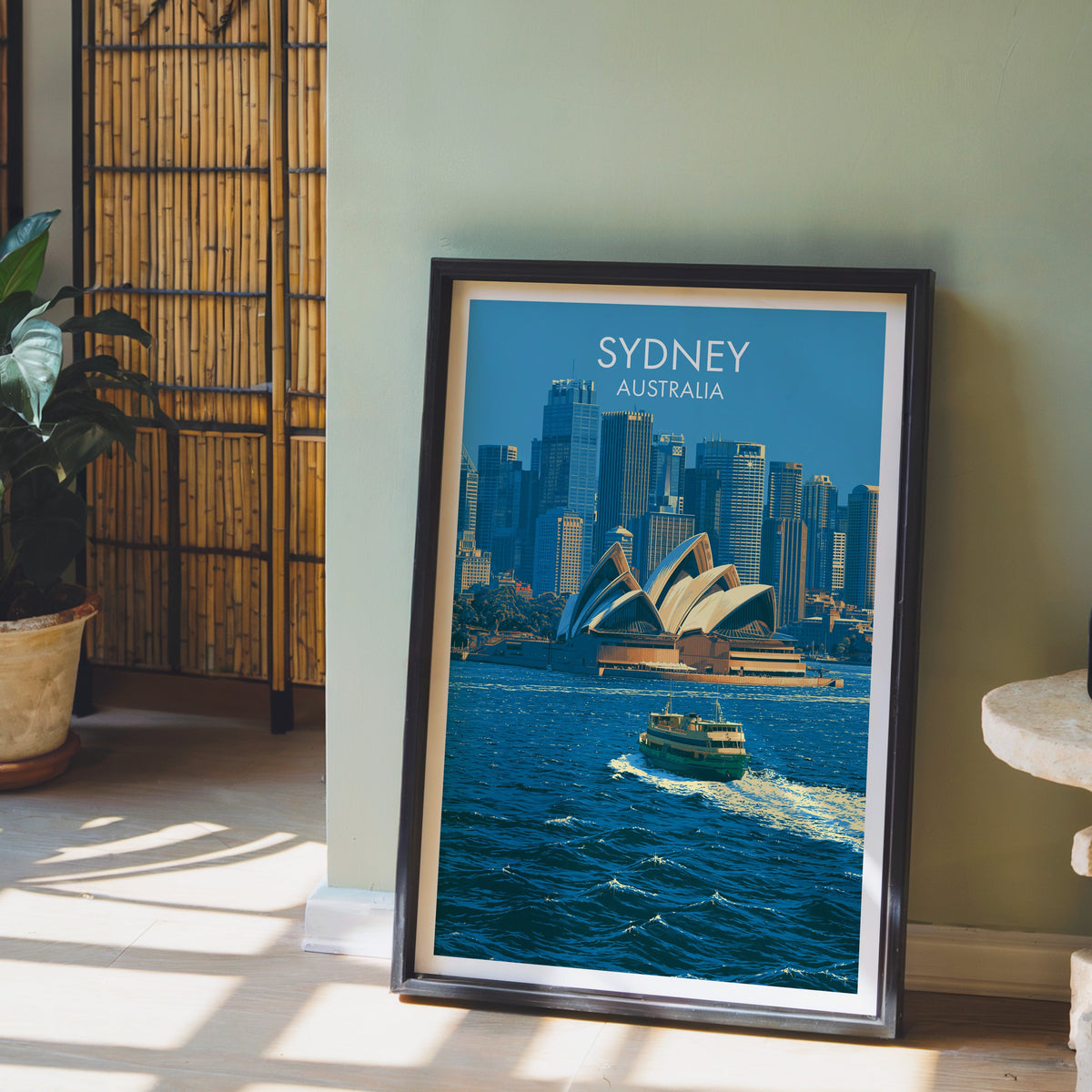 Sydney skyline artwork in a black frame on pale wood floor by a sage-green wall and bamboo screen