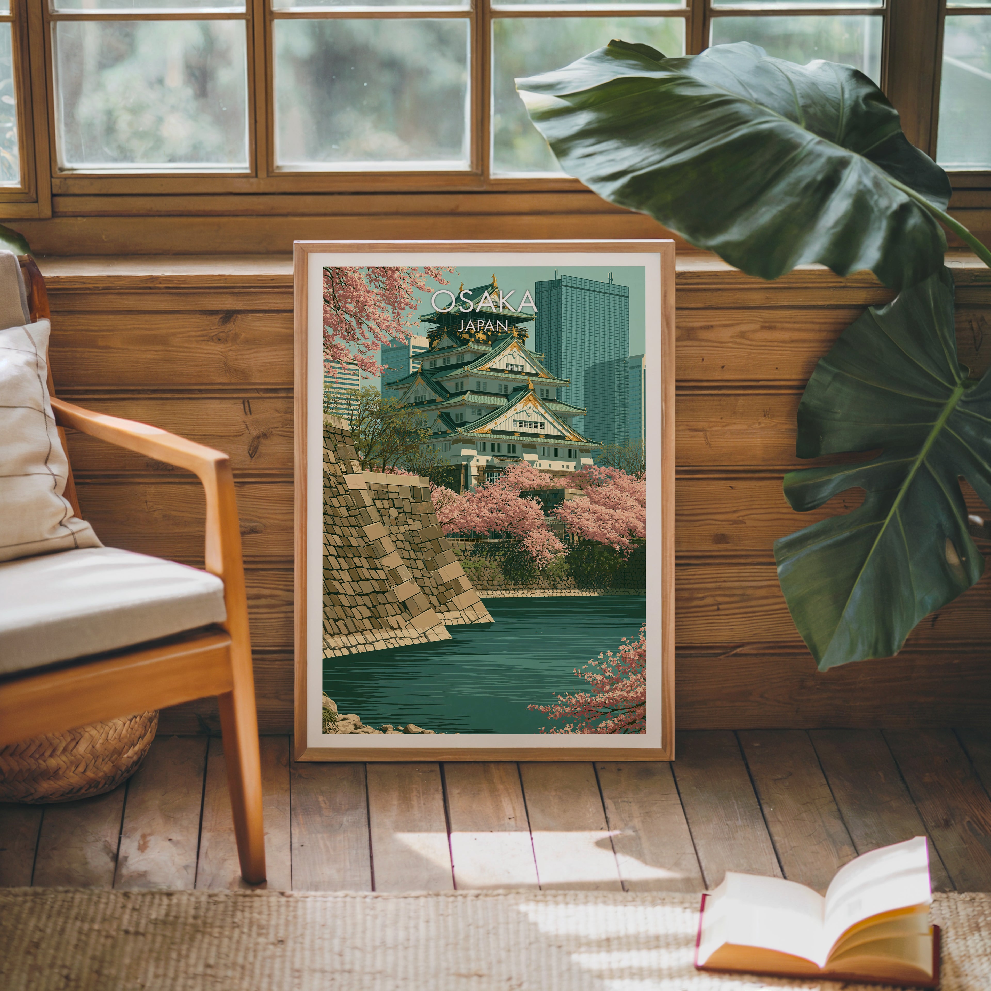 Framed Osaka Castle Japan poster leaning against a timber cabin wall with monstera leaves and an open book