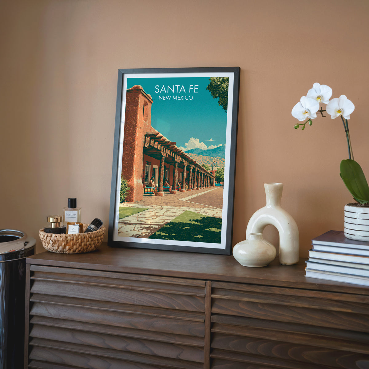 Santa Fe wall art in a black frame on a walnut sideboard by a terracotta wall with a cream vase and books.