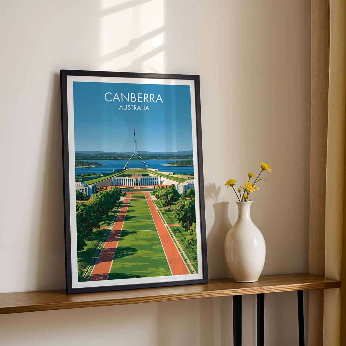 Parliament House artwork in a black frame on a wood shelf with yellow daisies on a sunlit cream wall