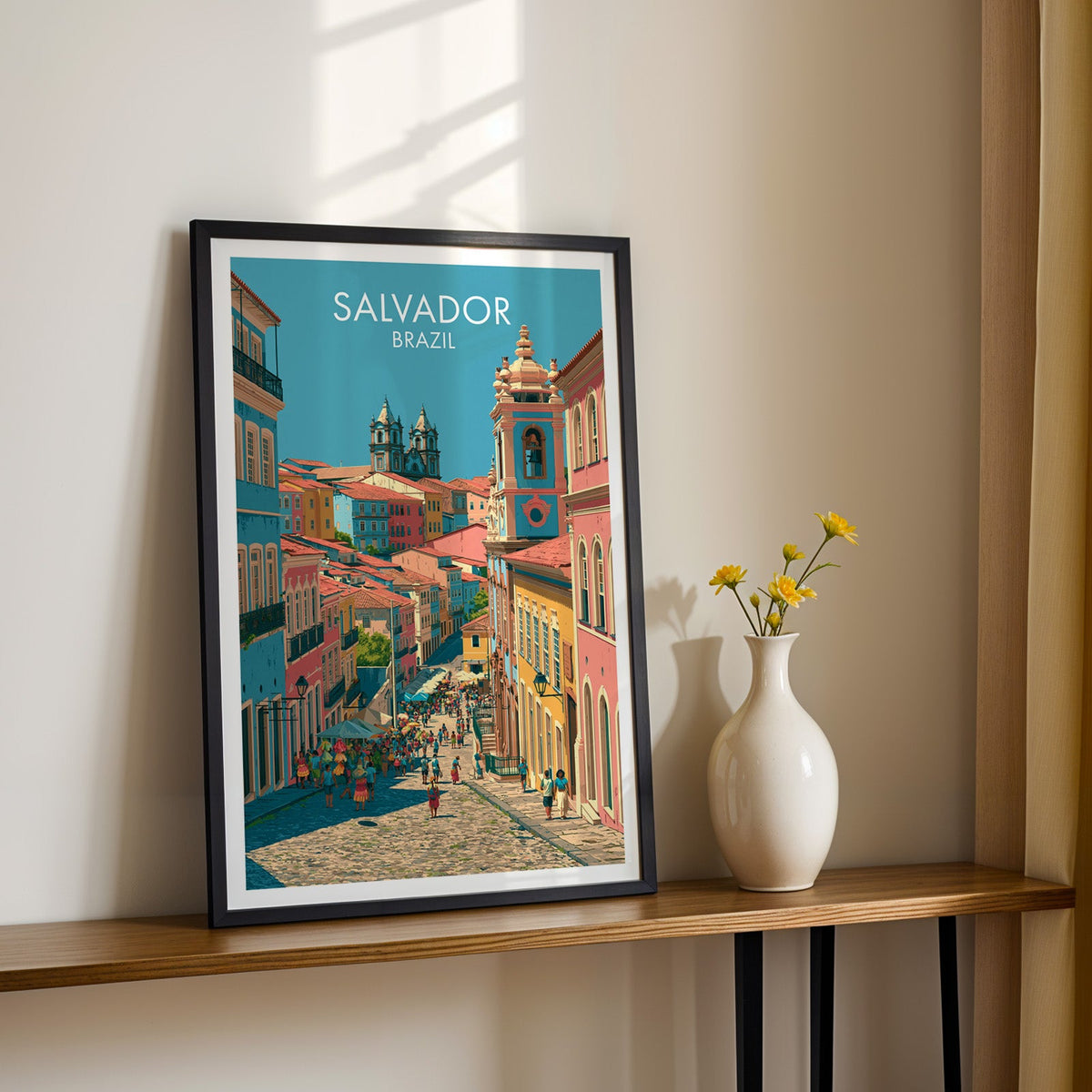 Cobbled street wall art in a black frame on a slim wood shelf with yellow daisies in a white vase, sunlit cream wall