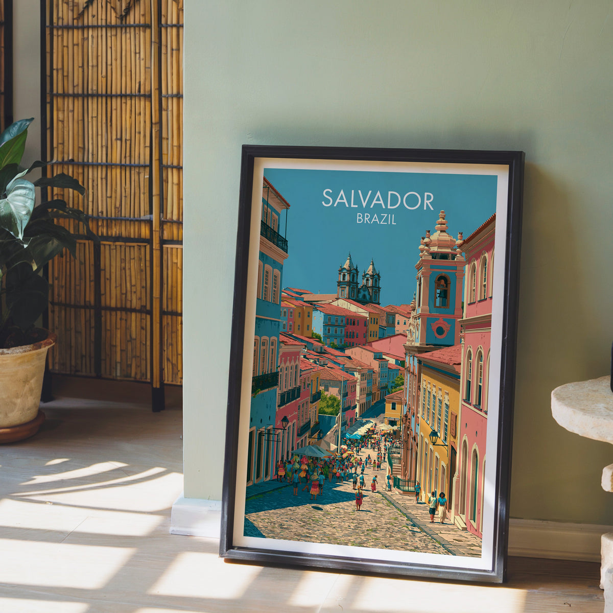 Bahia bell tower print in a dark brown frame on a pale wood floor by a sage wall, bamboo screen and terracotta pot