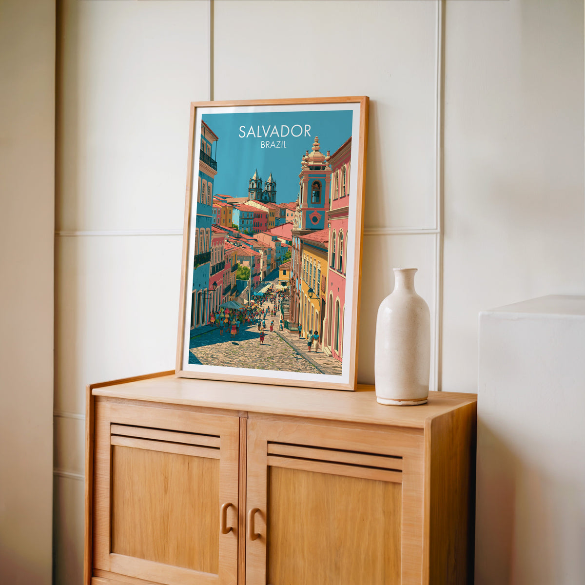 Coral townhouse art in an oak frame on a light oak cabinet by a cream panelled wall and a tall white vase