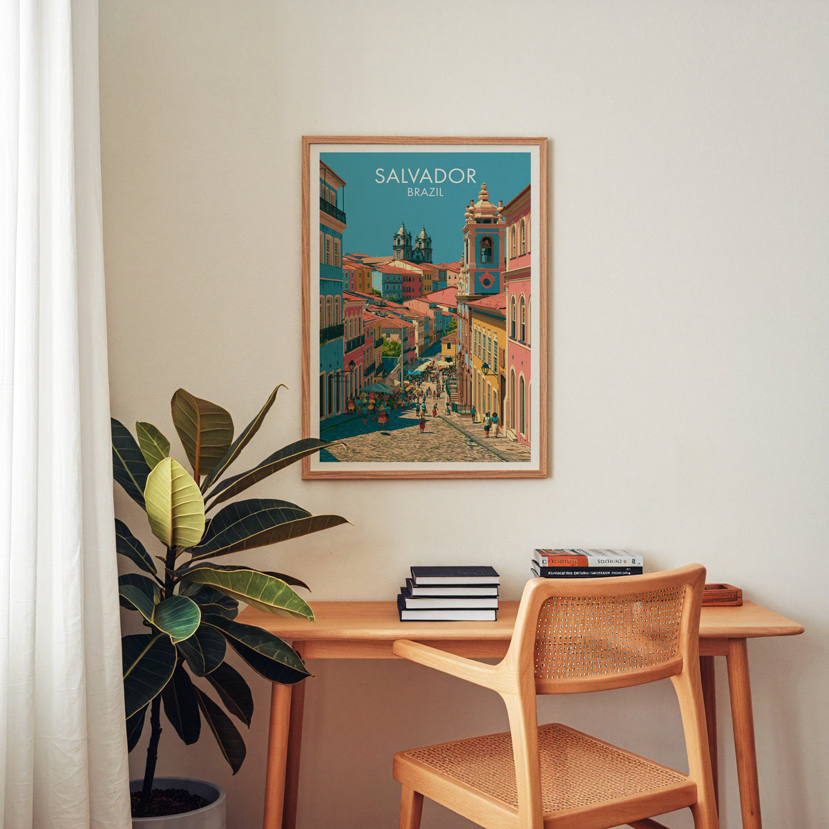 Pelourinho travel art in an oak frame over a wood desk with a cane chair, stacked books and a rubber plant