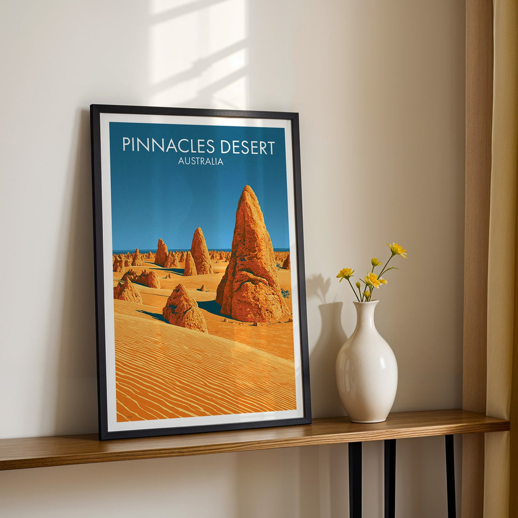 Golden desert artwork in a black frame on a wood shelf with yellow daisies on a sunlit cream wall