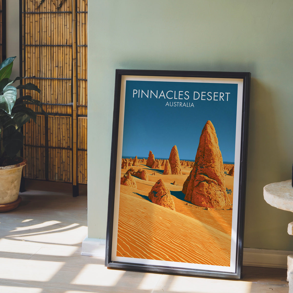 Orange dune and spire illustration in a black frame on pale wood floor by a sage wall and bamboo screen