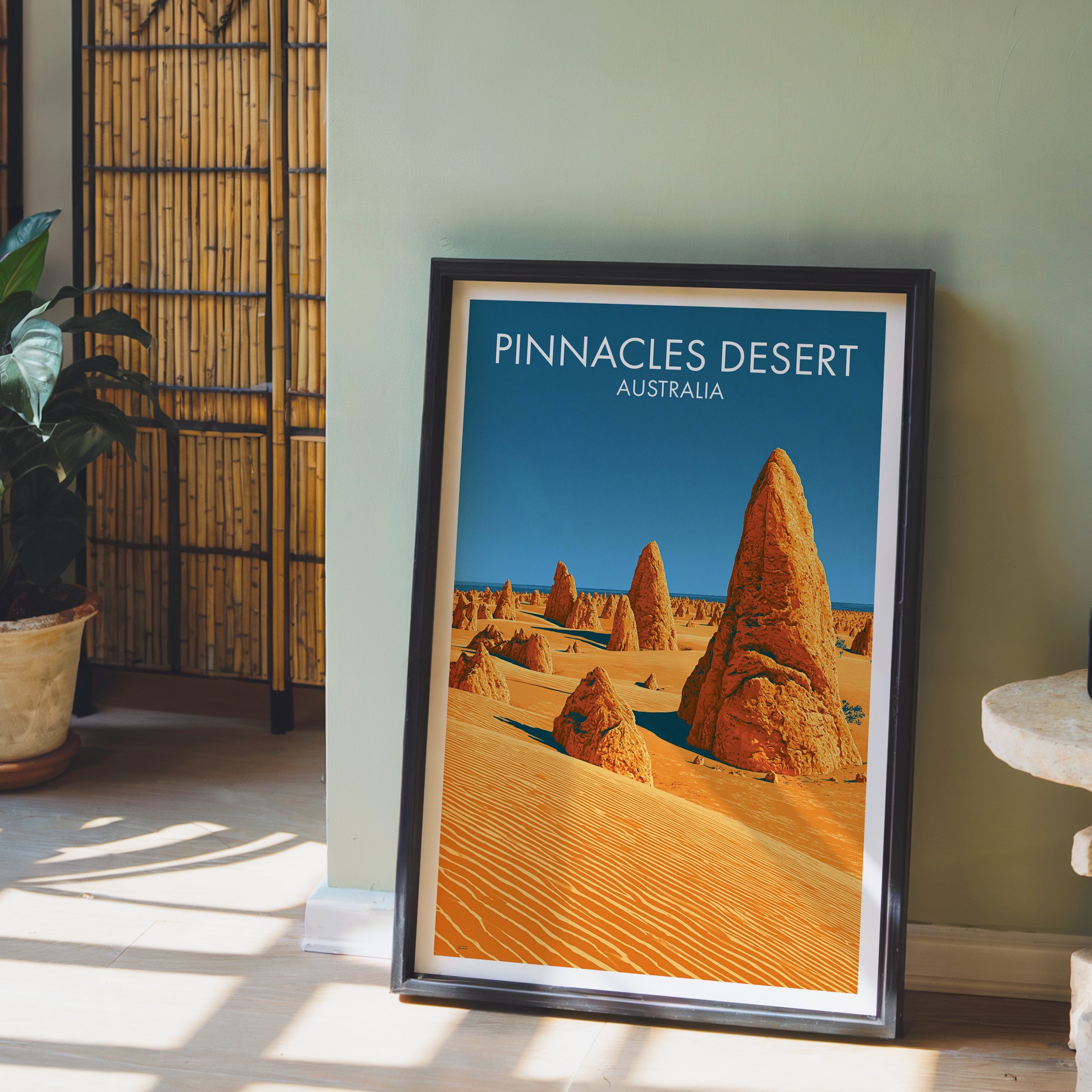 Orange dune and spire illustration in a black frame on pale wood floor by a sage wall and bamboo screen