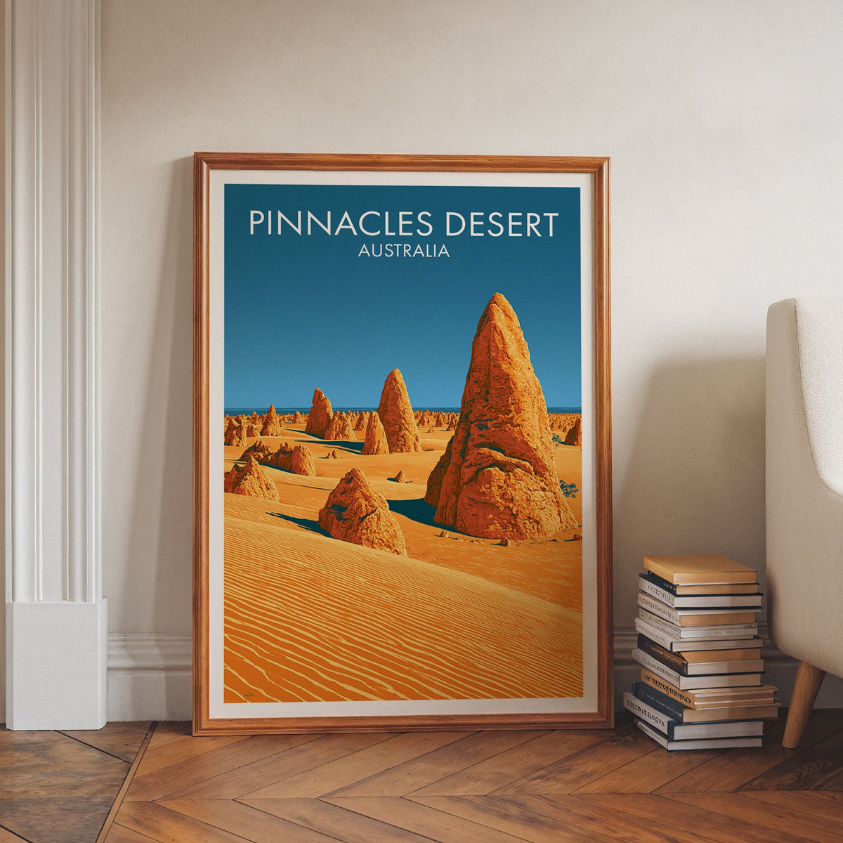 Rippled dune poster in a walnut frame on herringbone floor beside book stacks and a pale armchair