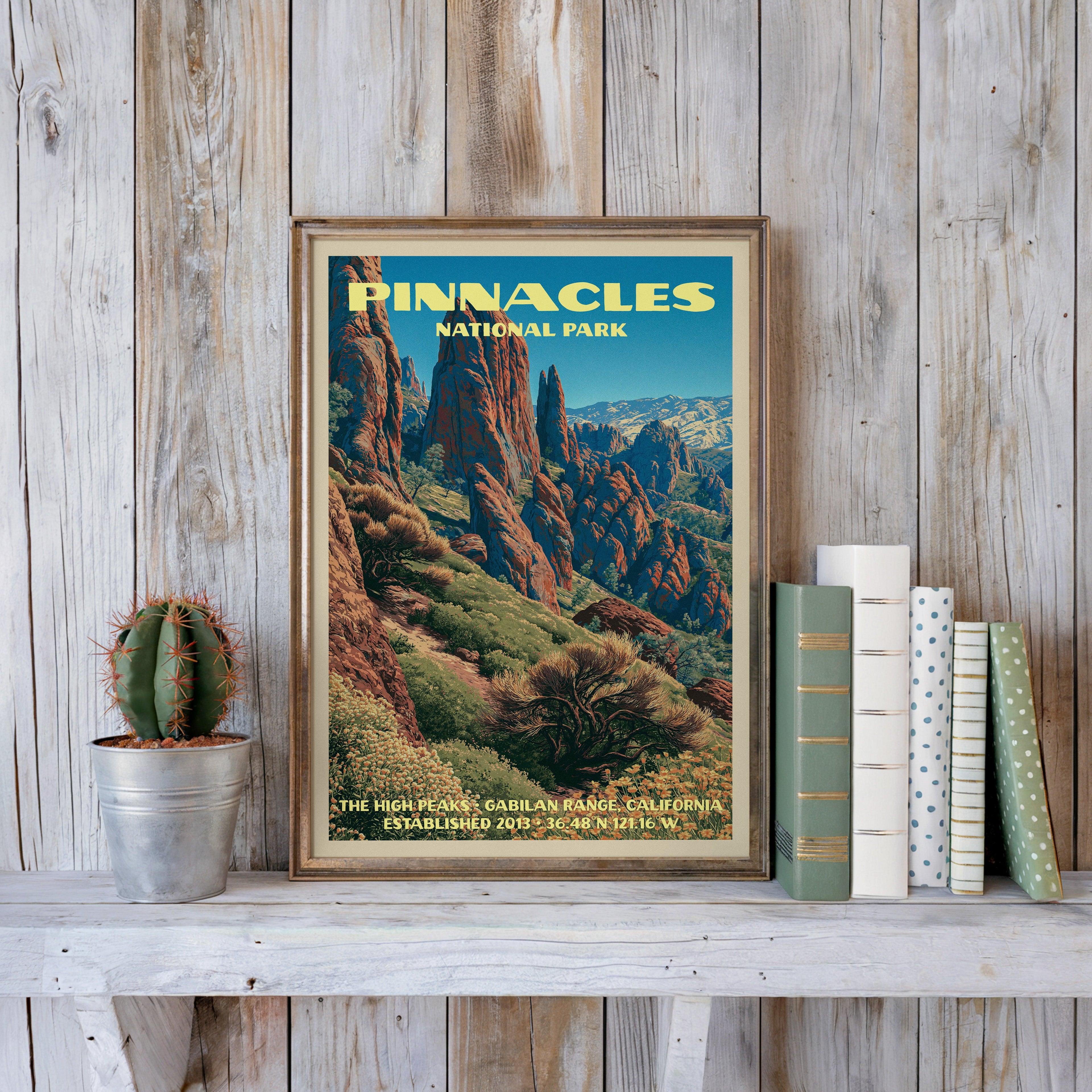 Pinnacles National Park poster in a slim gold frame on a whitewashed plank wall beside a cactus and pastel books