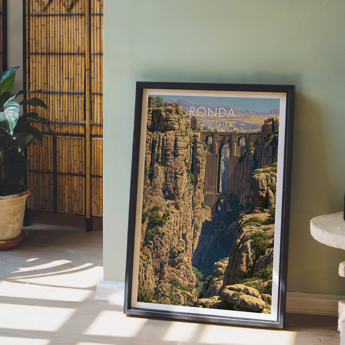 Puente Nuevo poster, dark frame leaning on a sage-green wall beside a bamboo screen and a fern