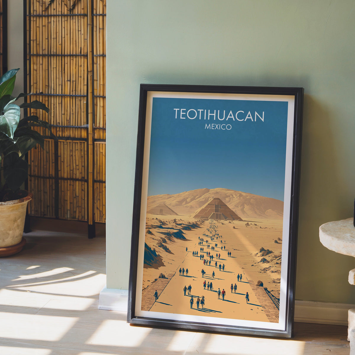 Sand and blue desert print in a black frame leaning on pale wood floor by a sage-green wall and bamboo screen