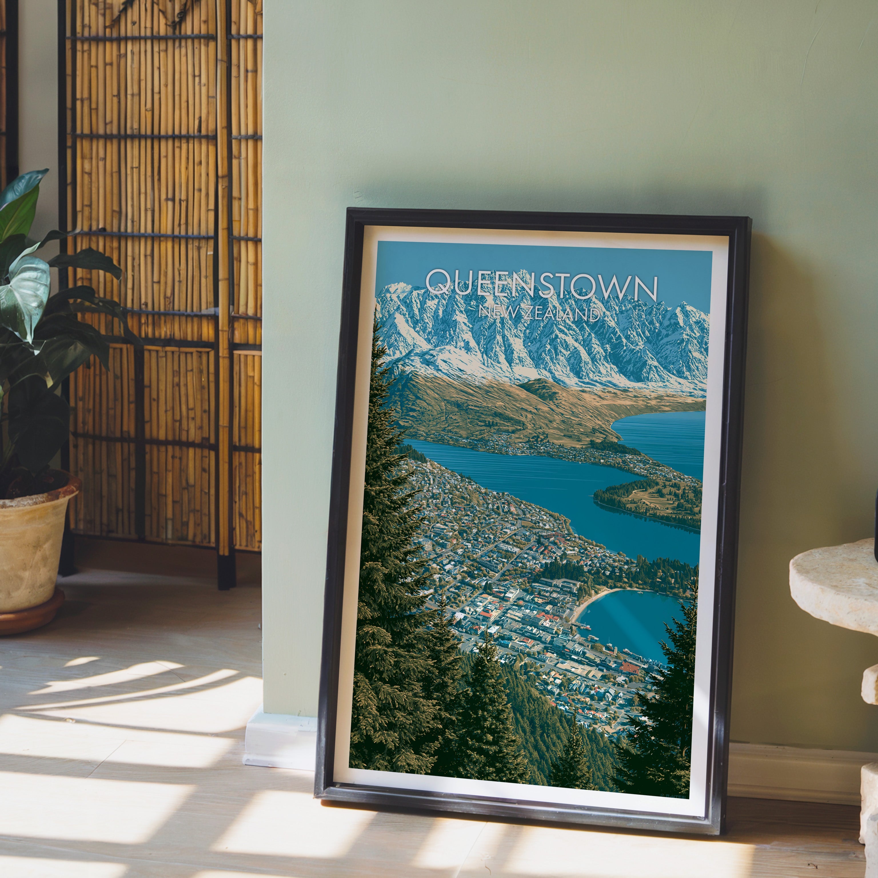 Alpine town print in a black frame on pale wood flooring by a sage-green wall and bamboo screen