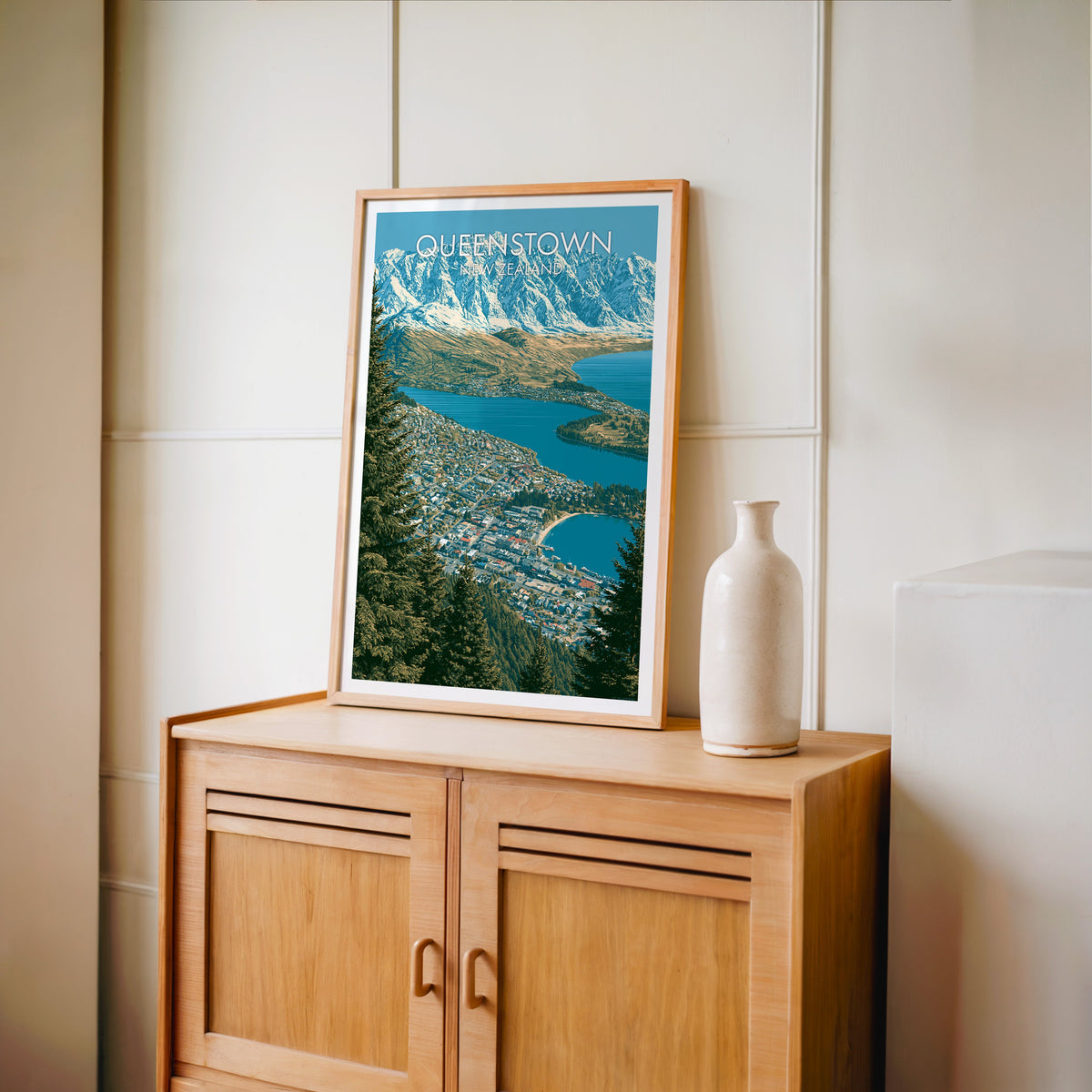 South Island town poster in an oak frame on a light oak cabinet beside a pale bottle vase