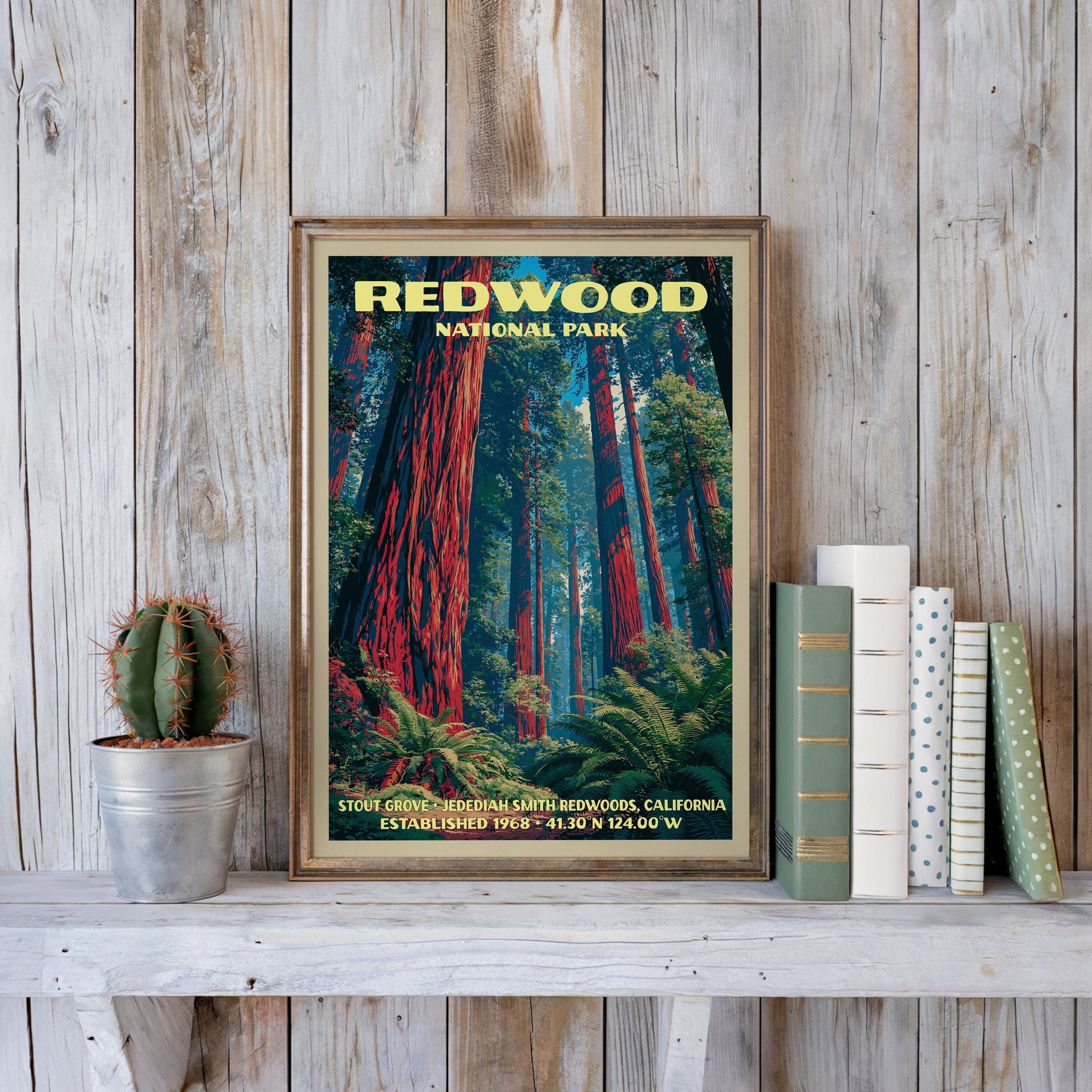 Redwood forest print in a slim gold frame on a whitewashed plank wall beside a cactus and pastel books