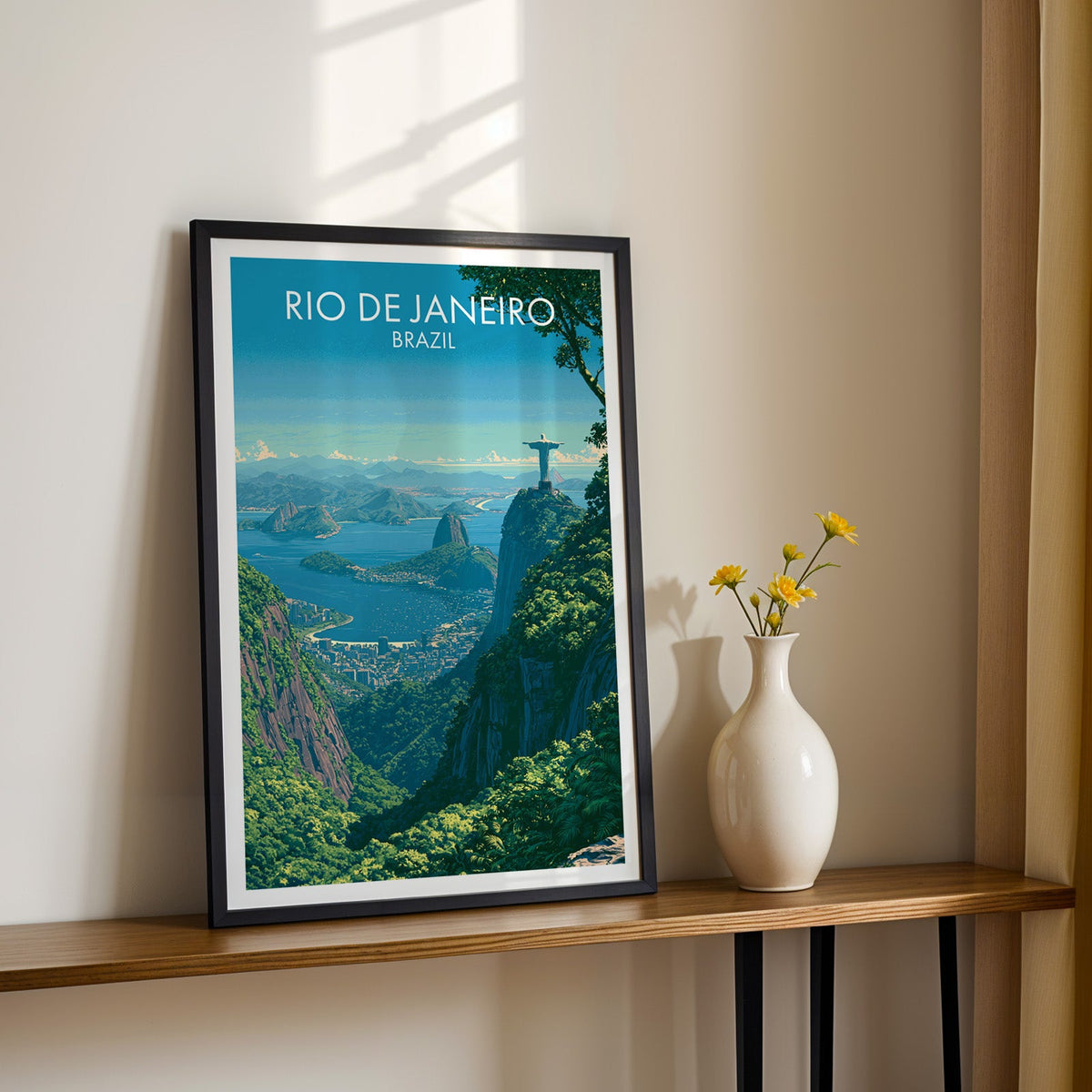 Guanabara Bay wall art in a black frame on a slim wood shelf beside yellow daisies in a white vase, sunlit cream wall