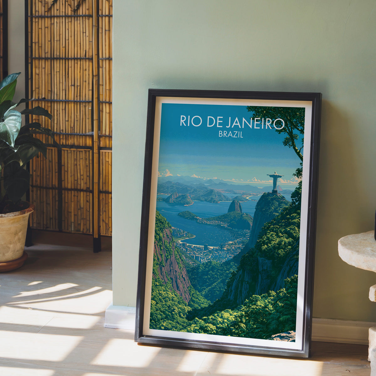 Corcovado overlook print in a dark brown frame on a pale wood floor by a sage wall, bamboo screen and terracotta pot