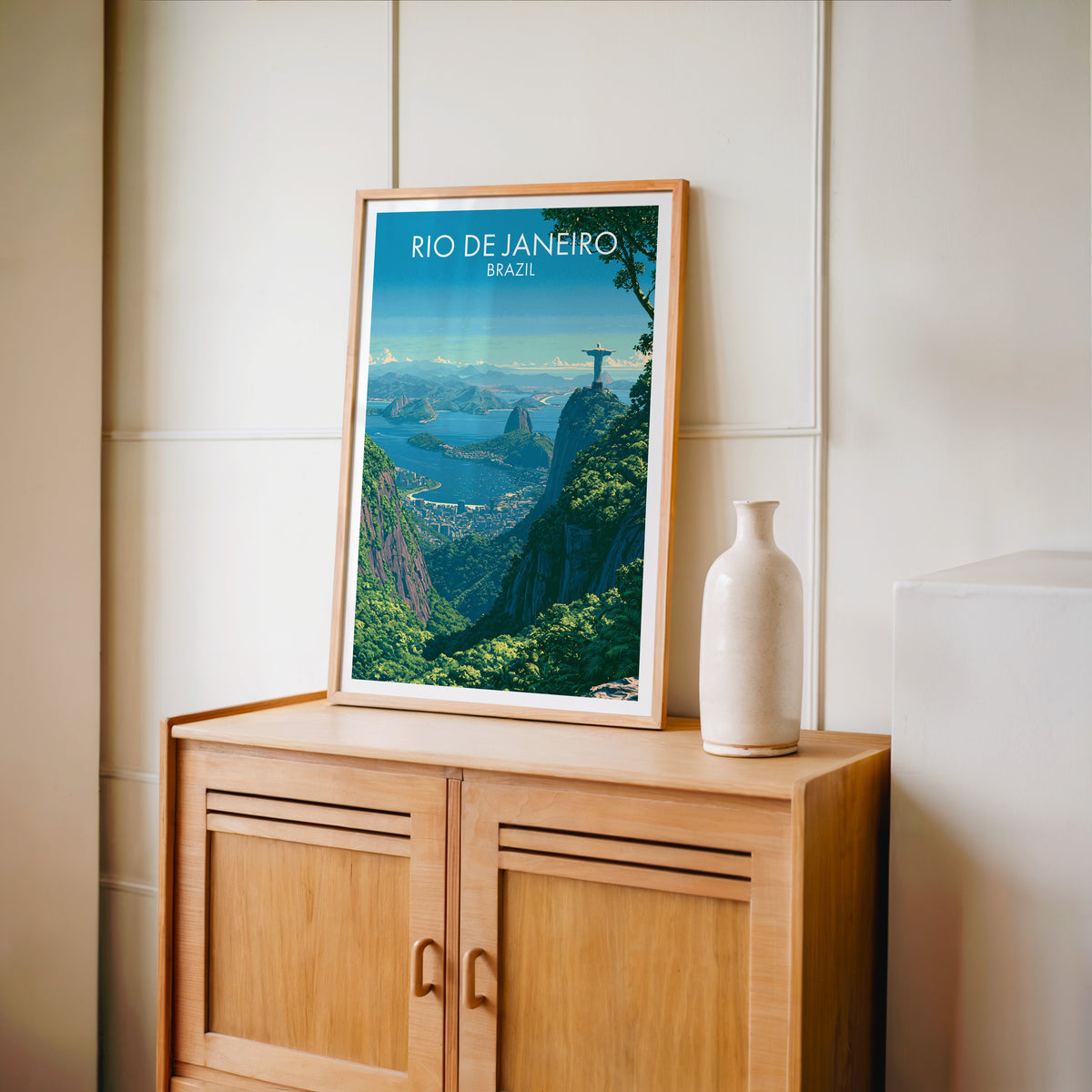 Christ the Redeemer travel art in an oak frame on a light oak cabinet by a cream panelled wall and tall white vase