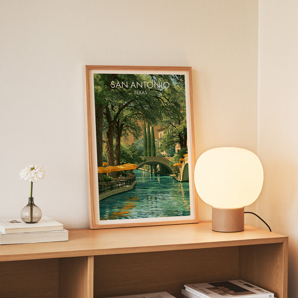 Green canal and bridge print in a slim oak frame on a console with daisies in a glass jar